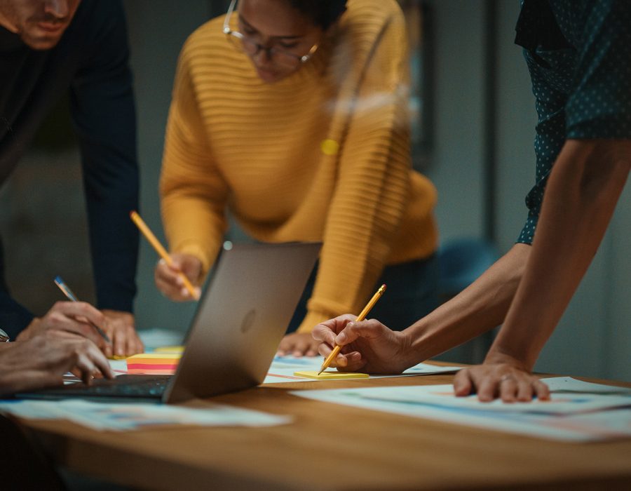 Close Up of Diverse Multiethnic Team Having Conversation in Meeting Room in a Creative Office. Colleagues Lean On a Conference Table, Look at Laptop Computer and Make Notes with Pencils on Notebooks.